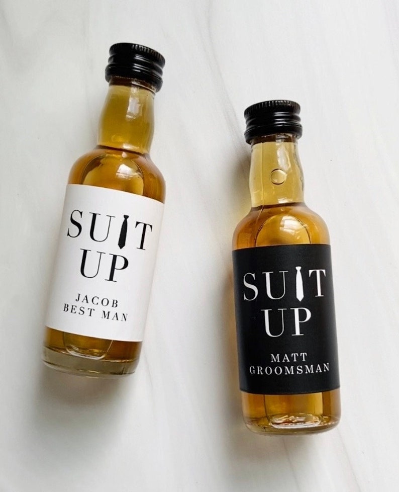 Puede incluir: Dos botellas peque&ntilde;as de licor con etiquetas blancas y negras que dicen "Suit Up". Una etiqueta tiene el texto "Jacob Best Man" y la otra etiqueta tiene el texto "Matt Groomsman".