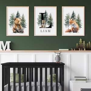 May include: Three framed watercolor prints featuring a bear, a fox, and a letter L with the name Liam. The prints are hung on a green wall above a black crib with a white star patterned sheet.