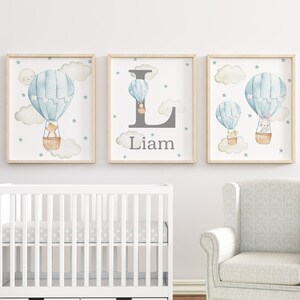 Puede incluir: Tres láminas enmarcadas para guardería con globos aerostáticos de acuarela en azul claro y beige. Una lámina tiene la letra "L" y el nombre "Liam". Las láminas están decoradas con nubes, estrellas y una luna.
