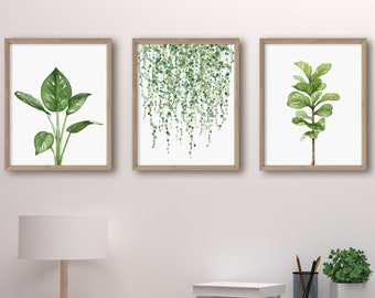 Könnte beinhalten: Drei gerahmte Aquarellbilder von grünen Pflanzen an einer weißen Wand über einem Holztisch mit einer Lampe, Büchern und einem Stiftehalter. Die Bilder zeigen eine Blattpflanze, eine hängende Rebe und einen Ficusbaum.