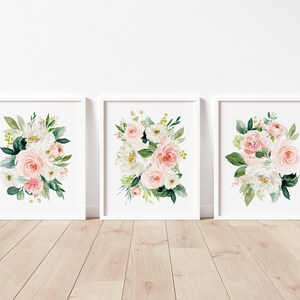 May include: Set of three floral prints featuring watercolor illustrations of pink and white roses, greenery, and white flowers. The prints are framed in white frames and displayed on a white wall.