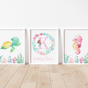 May include: Three watercolor prints featuring a sea turtle, a mermaid in a wreath with the letter K, and a seahorse. The prints are framed in white and have the name "Kingsley Grace" written below the mermaid.