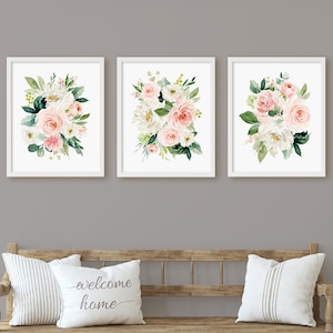 May include: Three framed floral prints with white frames and white mats. Each print features a watercolor painting of a bouquet of pink and white flowers with green leaves. The prints are hung on a gray wall above a wooden bench with two white pillows. One pillow has the text "welcome home" printed on it.
