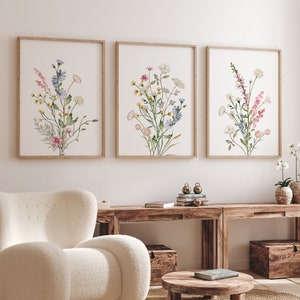 May include: Three framed floral watercolor prints. Each print features a different arrangement of wildflowers in shades of pink, blue, yellow, and green. The frames are a light brown color. The prints are displayed above a wooden console table.