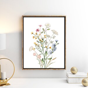 May include: A framed watercolor print of wildflowers. The artwork features a bouquet of delicate flowers in shades of pink, yellow, white, and blue, with green stems and leaves. The print is set against a white background and framed in a gold-toned frame.