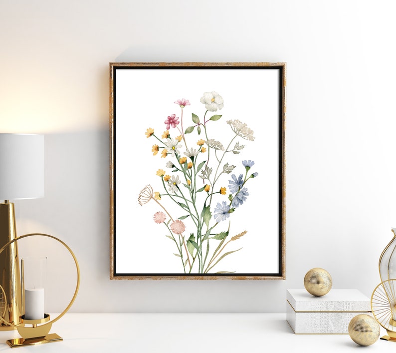 May include: A framed watercolor print of wildflowers. The artwork features a bouquet of delicate flowers in shades of pink, yellow, white, and blue, with green stems and leaves. The print is set against a white background and framed in a gold-toned frame.