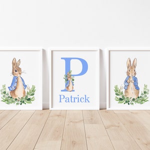May include: Three framed prints featuring a watercolor illustration of Peter Rabbit. The center print has the letter "P" in blue and the name "Patrick" below it. The prints are on a light wood floor.