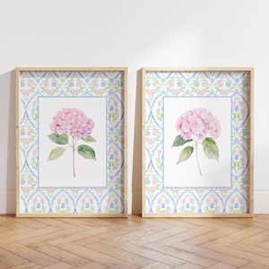 May include: Two framed watercolor prints featuring pink hydrangea flowers. Each print has a light blue and floral patterned border, set within a light wood frame. The prints are displayed on a wooden floor against a white wall.