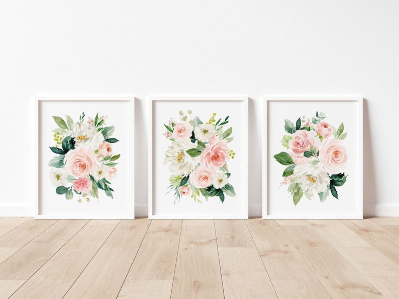 May include: Set of three floral prints featuring watercolor illustrations of pink and white roses, greenery, and white flowers. The prints are framed in white frames and displayed on a white wall.