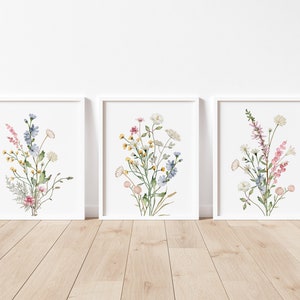 May include: Three framed watercolor prints featuring bouquets of wildflowers. Each print showcases a different arrangement of delicate blooms in shades of pink, yellow, blue, and white, set against a white background. The frames are white, and the artwork is displayed on a wooden floor.