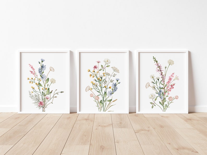 May include: Three framed watercolor prints featuring bouquets of wildflowers. Each print showcases a different arrangement of delicate blooms in shades of pink, yellow, blue, and white, set against a white background. The frames are white, and the artwork is displayed on a wooden floor.