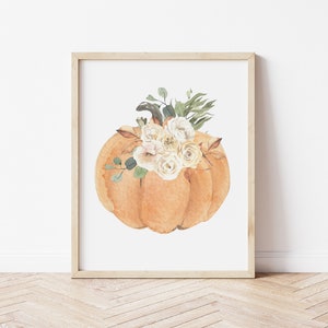 May include: A watercolor painting of an orange pumpkin with white flowers on top. The pumpkin is in a simple, white frame.