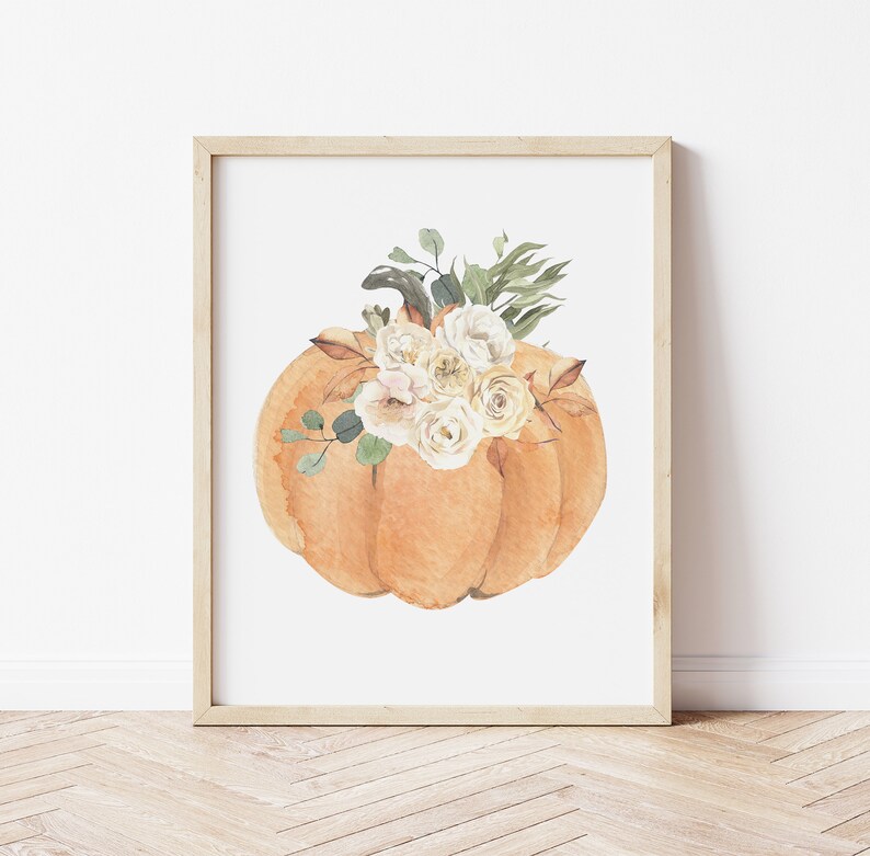 May include: A watercolor painting of an orange pumpkin with white flowers on top. The pumpkin is in a simple, white frame.