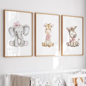 May include: Three framed watercolor prints featuring adorable baby animals: an elephant, a giraffe, and a zebra. Each animal is depicted in a seated position and adorned with a pink bow. The prints have a light wood frame and are displayed on a white wall.