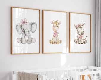 May include: Three framed watercolor prints featuring adorable baby animals: an elephant, a giraffe, and a zebra. Each animal is depicted in a seated position and adorned with a pink bow. The prints have a light wood frame and are displayed on a white wall.