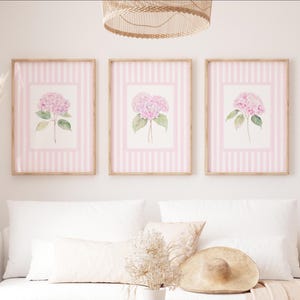 May include: Three framed floral prints featuring pink hydrangea blooms with green leaves. The artwork is set against a pink and white striped background, encased in light brown wooden frames. The overall aesthetic is soft and feminine.