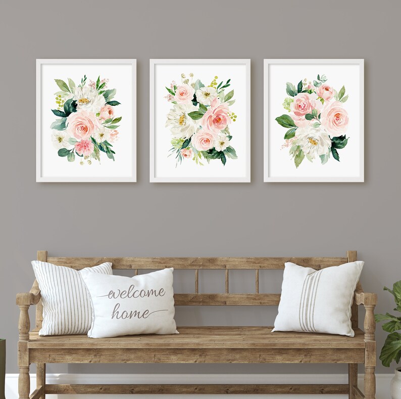 May include: Three framed floral prints with white frames and white mats. Each print features a watercolor painting of a bouquet of pink and white flowers with green leaves. The prints are hung on a gray wall above a wooden bench with two white pillows. One pillow has the text "welcome home" printed on it.
