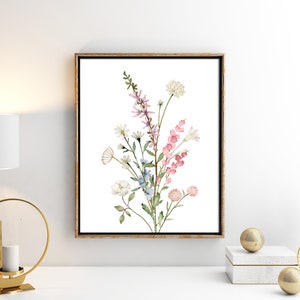 May include: A framed watercolor print featuring a bouquet of wildflowers in shades of pink, white, and blue. The artwork is set against a white background and framed in a gold-toned wooden frame. The floral arrangement includes daisies, snapdragons, and other delicate blooms.