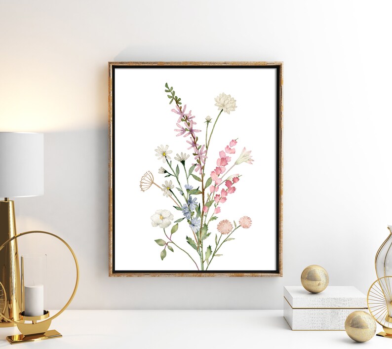 May include: A framed watercolor print featuring a bouquet of wildflowers in shades of pink, white, and blue. The artwork is set against a white background and framed in a gold-toned wooden frame. The floral arrangement includes daisies, snapdragons, and other delicate blooms.