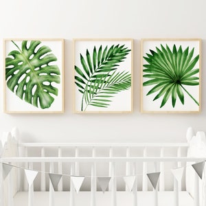 K&ouml;nnte beinhalten: Drei gerahmte Drucke von Aquarell-Illustrationen von tropischen Bl&auml;ttern. Die Drucke sind in Holzrahmen gefasst und an einer wei&szlig;en Wand &uuml;ber einem wei&szlig;en Kinderbett mit einer silbernen Girlande aufgeh&auml;ngt.