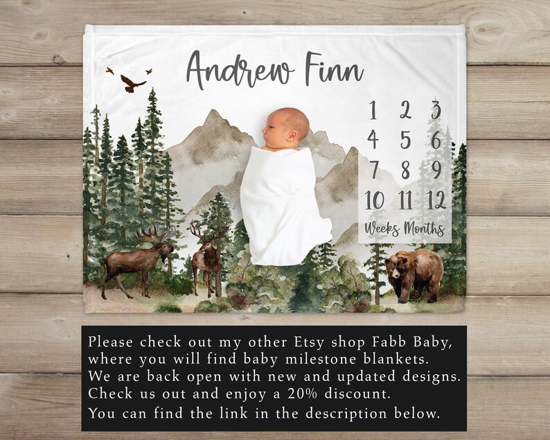 May include: A white baby blanket with a watercolor forest scene, mountains, and a baby bear. The blanket has a personalized name "Andrew Finn" and a number chart for tracking baby's growth in weeks and months.
