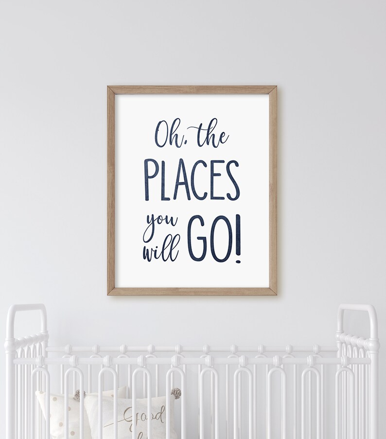 May include: A white framed print with the text "Oh, the PLACES you will GO!" in navy blue script. The print is hanging on a white wall above a white crib.