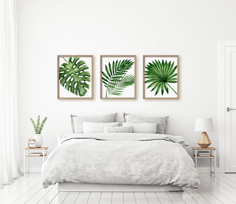 K&ouml;nnte beinhalten: Drei gerahmte Drucke von tropischen Bl&auml;ttern in einem Schlafzimmer. Die Drucke sind in Holzrahmen und zeigen ein Monstera-Blatt, eine Palmenwedel und ein F&auml;cherpalmenblatt. Die Drucke h&auml;ngen an einer wei&szlig;en Wand &uuml;ber einem grauen Bett mit wei&szlig;en Bettw&auml;sche.