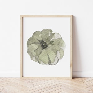 May include: A framed watercolor painting of a green pumpkin. The pumpkin is painted in a light green color with darker green accents. The painting is on a white background and is framed in a light wood frame.