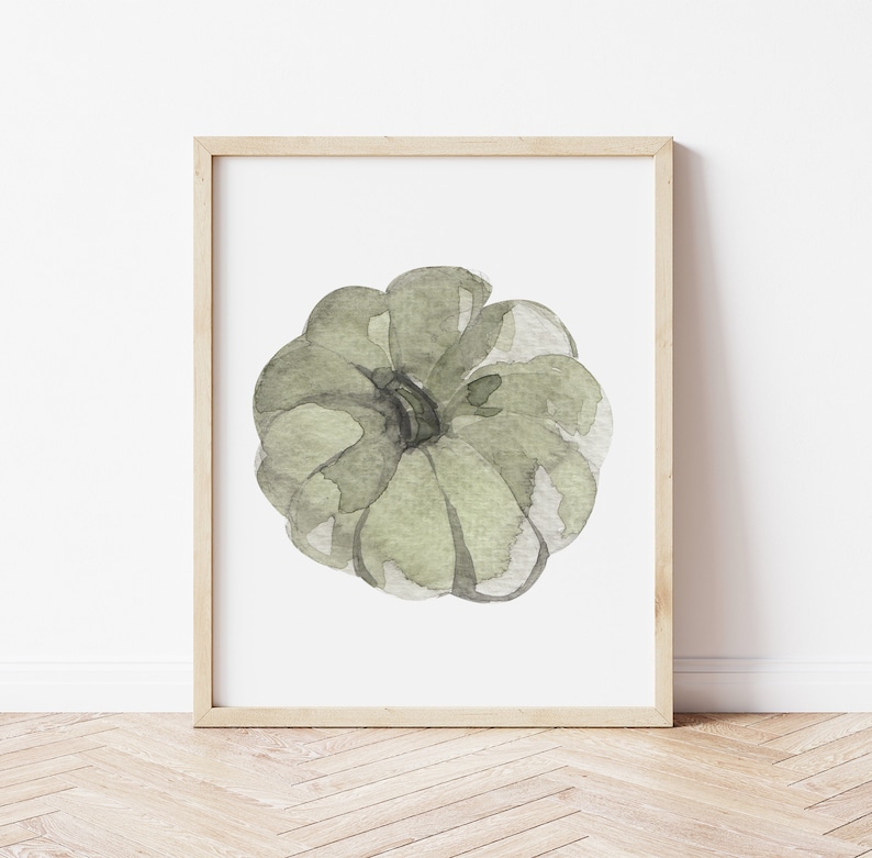May include: A framed watercolor painting of a green pumpkin. The pumpkin is painted in a light green color with darker green accents. The painting is on a white background and is framed in a light wood frame.