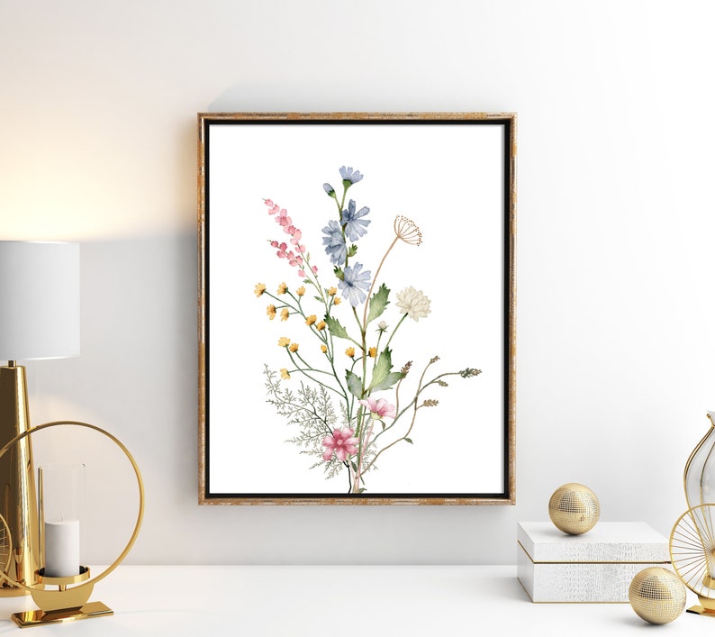 May include: A framed watercolor print featuring a bouquet of wildflowers. The artwork showcases pink, blue, yellow, and white flowers with green foliage against a white background. The frame is a warm gold color.