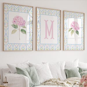 May include: Three framed wall art prints featuring a light blue and pink floral trellis border. The left and right prints display a single pink hydrangea flower with green leaves. The center print features a large, centered pink letter M.