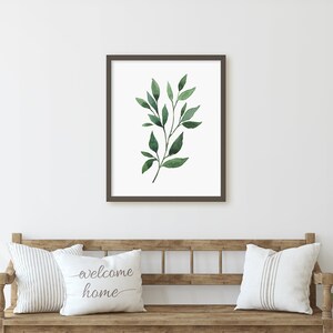 May include: A framed print of a watercolor painting of a green leafy branch. The print is hanging on a white wall above a wooden bench with two white pillows. One pillow has the text "welcome home" printed on it.