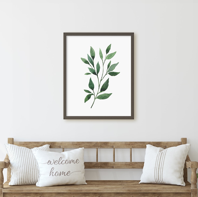 May include: A framed print of a watercolor painting of a green leafy branch. The print is hanging on a white wall above a wooden bench with two white pillows. One pillow has the text "welcome home" printed on it.
