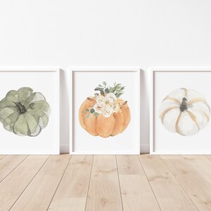 May include: Three watercolor paintings of pumpkins. The first pumpkin is green, the second pumpkin is orange with white flowers, and the third pumpkin is white with gold accents.