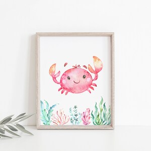 May include: A watercolour painting of a pink crab with a smiling face, surrounded by green and orange seaweed. The crab is in the centre of the image, and the seaweed is around the edges.