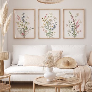 May include: Three framed botanical art prints featuring watercolor illustrations of wildflowers in shades of pink, blue, yellow, and green. The prints are displayed above a white sofa with decorative pillows, a round coffee table, and a woven lamp.