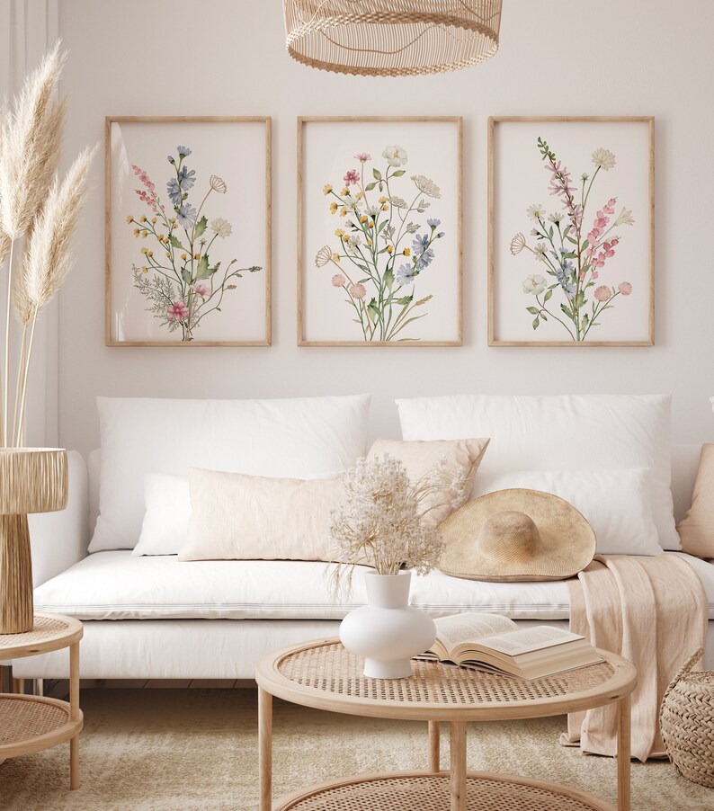 May include: Three framed botanical art prints featuring watercolor illustrations of wildflowers in shades of pink, blue, yellow, and green. The prints are displayed above a white sofa with decorative pillows, a round coffee table, and a woven lamp.