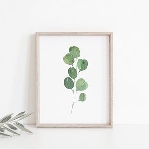 May include: A framed watercolor painting of a green eucalyptus branch with leaves. The painting is on a white background and is framed in a light wood frame.