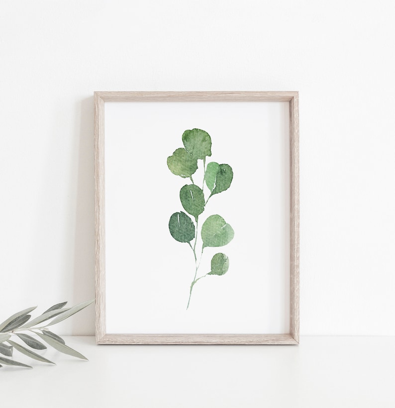 May include: A framed watercolor painting of a green eucalyptus branch with leaves. The painting is on a white background and is framed in a light wood frame.