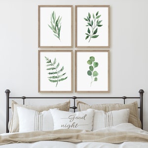 May include: Four framed watercolor prints of green leaves and branches. The prints are arranged in a grid pattern on a white wall above a bed with a tan headboard and pillows. The pillows have a white pillowcase with the text "Good night" printed on it.