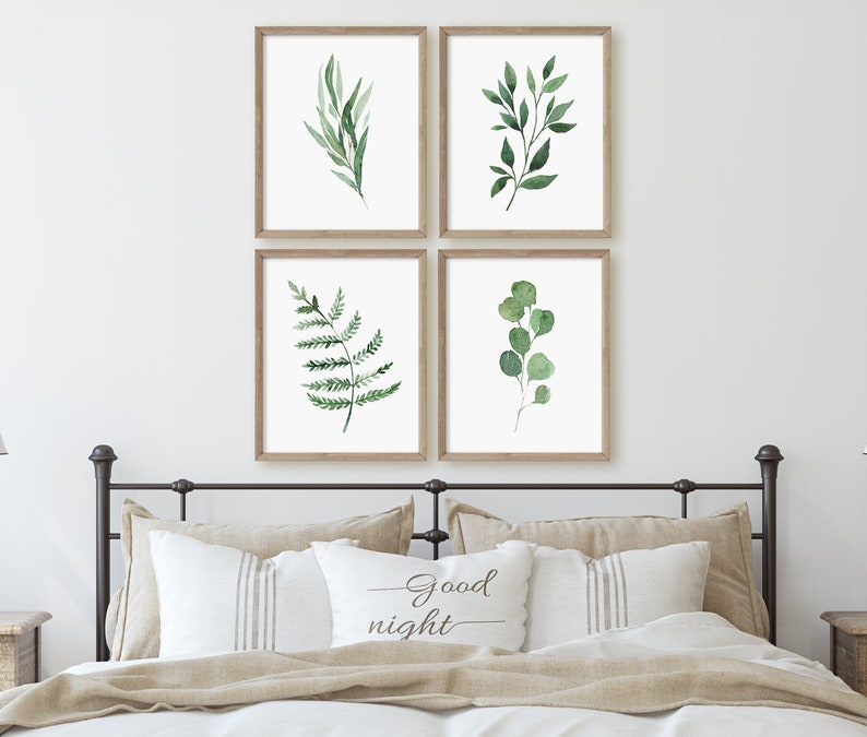 May include: Four framed watercolor prints of green leaves and branches. The prints are arranged in a grid pattern on a white wall above a bed with a tan headboard and pillows. The pillows have a white pillowcase with the text "Good night" printed on it.