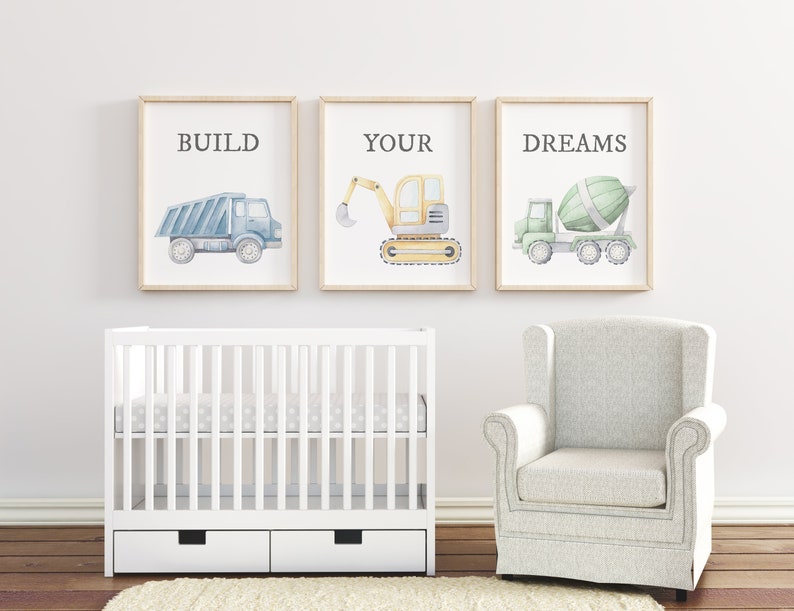 May include: Three framed watercolor prints with construction vehicles: a blue dump truck with the word "BUILD", a yellow excavator with "YOUR", and a green cement mixer with "DREAMS". The prints are above a white crib and a gray armchair.
