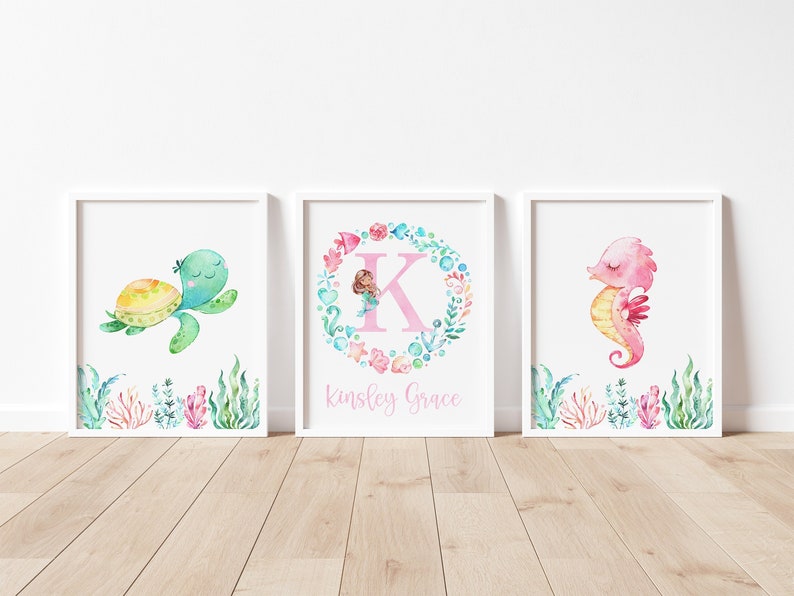 May include: Three watercolor prints featuring a sea turtle, a mermaid in a wreath with the letter K, and a seahorse. The prints are framed in white and have the name "Kingsley Grace" written below the mermaid.