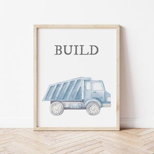 May include: A framed print featuring a watercolor illustration of a blue dump truck. The word "BUILD" is printed in gray above the truck. The artwork is set against a white background, framed in light wood.