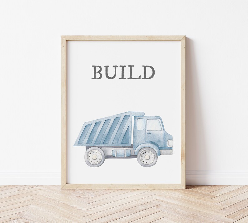 May include: A framed print featuring a watercolor illustration of a blue dump truck. The word "BUILD" is printed in gray above the truck. The artwork is set against a white background, framed in light wood.