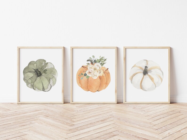 May include: Three framed watercolor paintings of pumpkins. The first pumpkin is green, the second is orange with white flowers, and the third is white with gold accents.