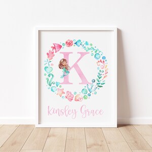 May include: A white framed print with a pink letter K surrounded by a wreath of watercolor sea-themed elements, including a mermaid, seashells, starfish, and anchors. The name Kinsley Grace is written below the letter in pink.