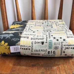 May include: A white and yellow dinosaur-themed baby blanket with black text that says "Big Dinosaur", "Enormous", "Bones", "Fossil", "Extinct", "Stomp", "Brachiosaurus", "Stegosaurus", and "Jurassic".