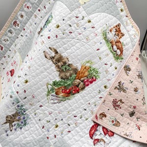 May include: A quilted blanket featuring a whimsical woodland theme. The design includes illustrations of a rabbit, fox, mushrooms, and flowers. The quilt has a white background with gray and pink accents, and is likely made of cotton.