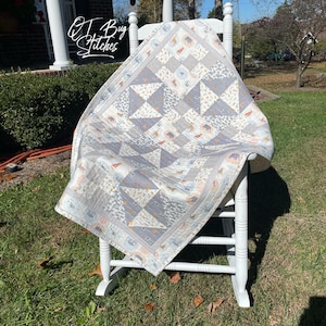 May include: A handmade quilt with a patchwork design, draped over a white rocking chair. The quilt features a pattern of gray, white, and tan triangles and squares. The quilt has a border with animal and floral motifs. The text "QT Bug Stitches" is visible.